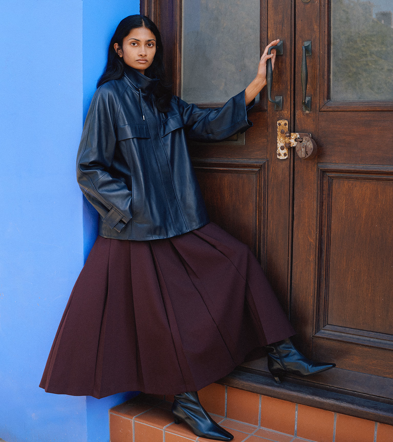 Model against a blue wall in a navy leather funnel-neck anorak, plum pleated kilt skirt and black knee-high boots