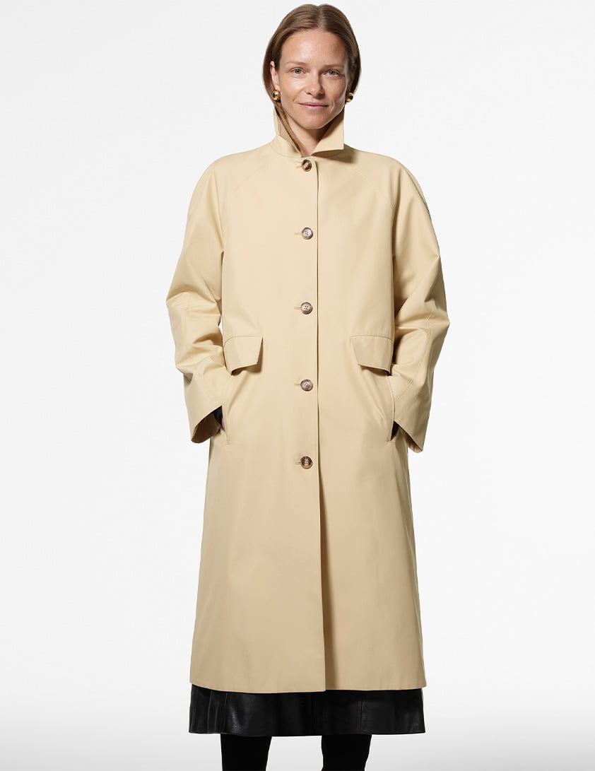 Woman wearing Whistles' long beige coat with a collar, button front and flap pockets, layered over black leather skirt