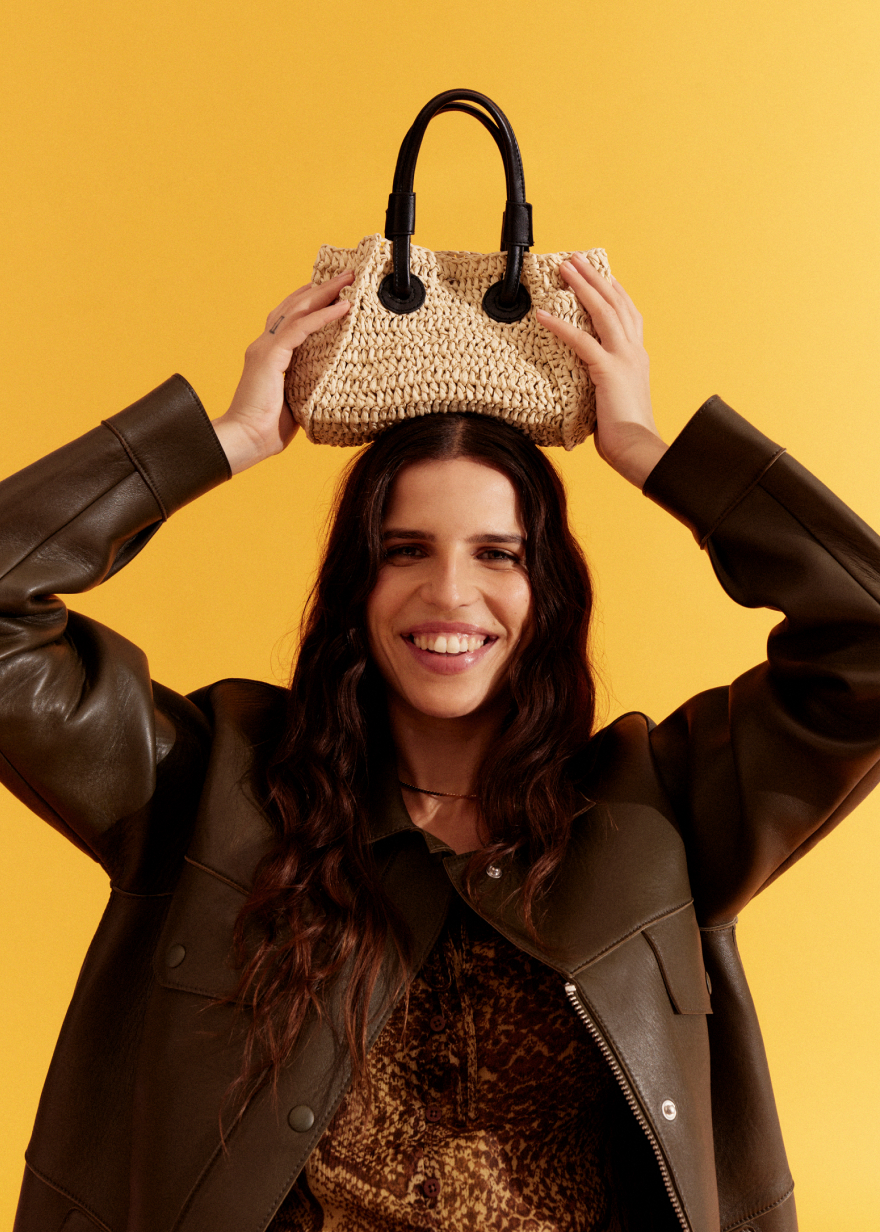 Smiling model in a dark brown leather jacket holding a raffia tote above her head against a yellow background.
