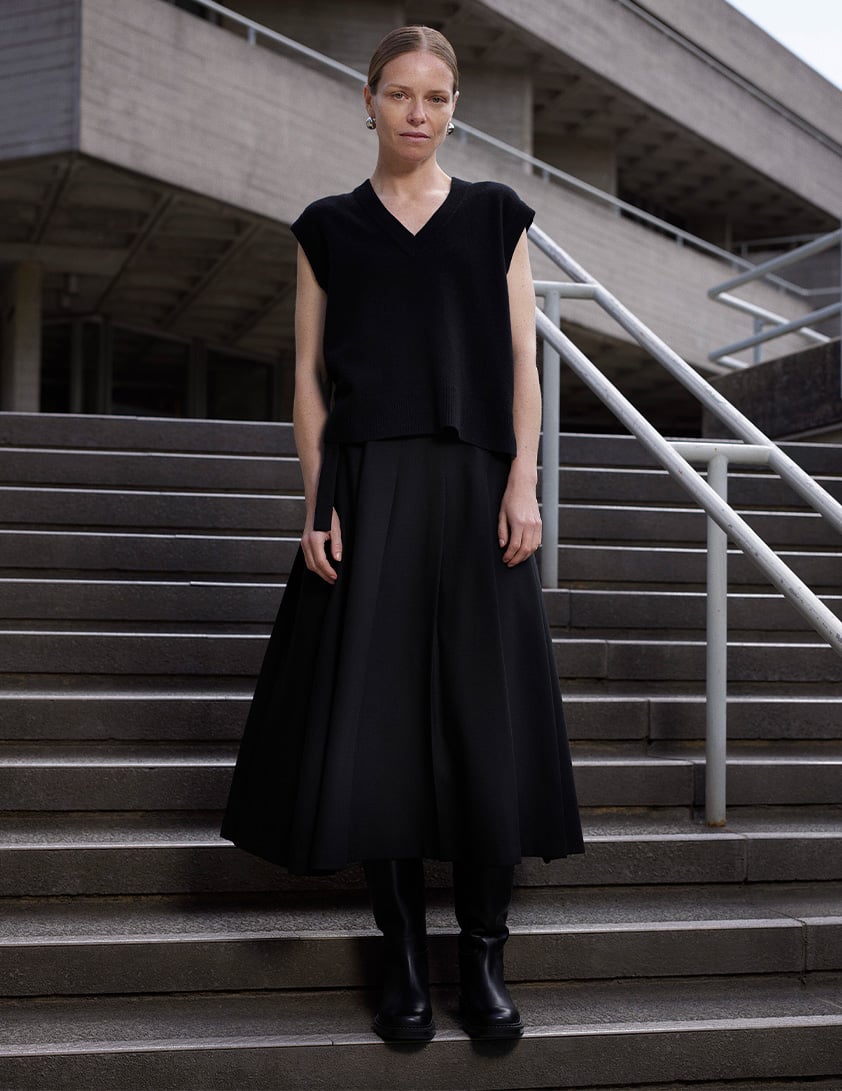 Woman wearing Whistles' black sleeveless vest with a flared midi skirt and black knee-high leather boots, styled with silver earrings