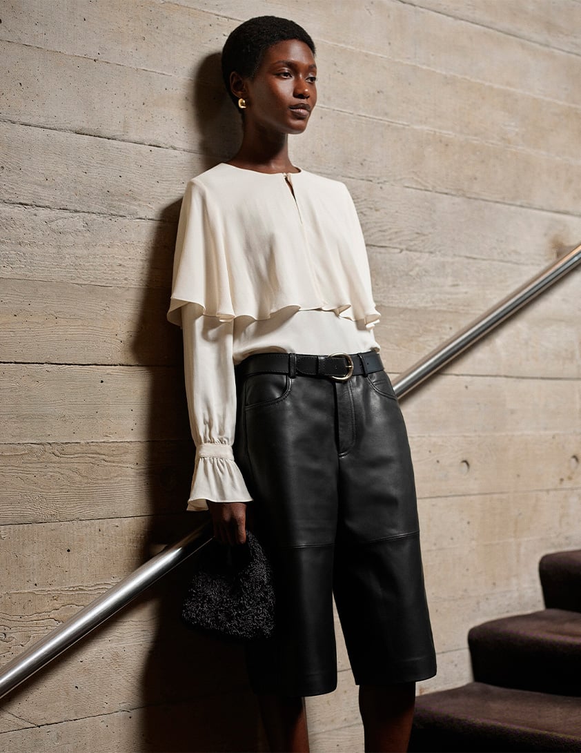 Woman wearing Whistles' cream layered blouse with flared cuff sleeves, black leather knee-length shorts with belt, holding textured black handbag