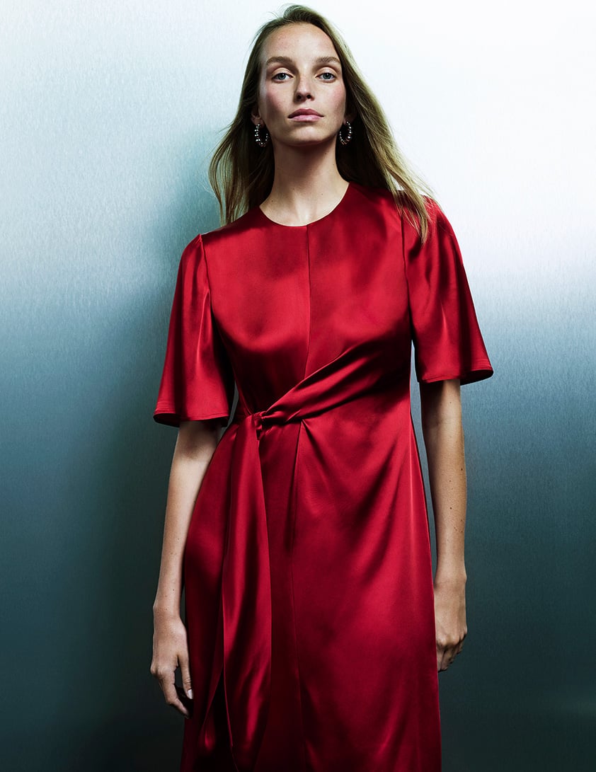Woman wearing a Whistles red satin dress with short flutter sleeves and tie knot waist detail.