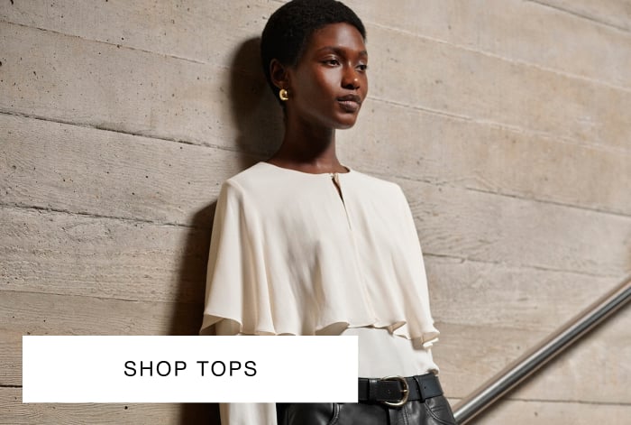 Woman wearing Whistles' cream layered blouse with flared cuff sleeves, black leather knee-length shorts with belt, holding textured black handbag