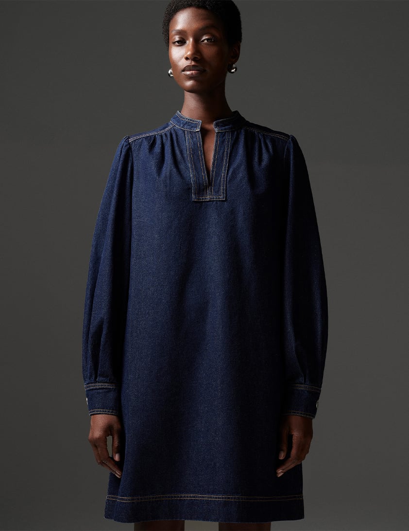Woman wearing Whistles' indigo denim midi dress with split neckline, long sleeves and contrast stitching, styled with silver earrings