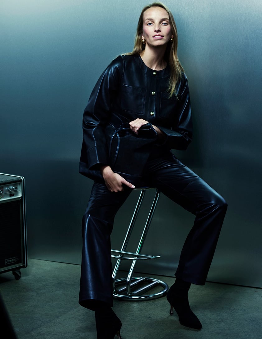 Woman wearing a Whistles black leather jacket with gold button detailing and matching pants, holding a black leather handbag