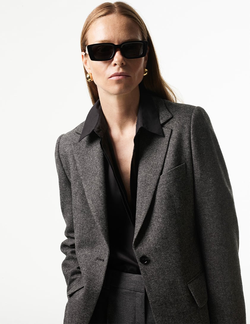 Woman wearing Whistles tailored grey blazer with black shirt gold hoop earrings and black sunglasses now 20% off.