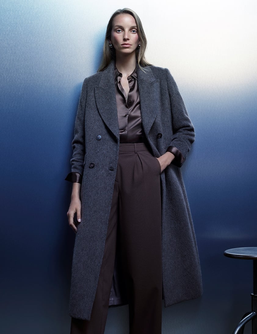 Woman wearing a Whistles grey double-breasted long coat with a brown satin shirt tucked into high waisted tailored trousers