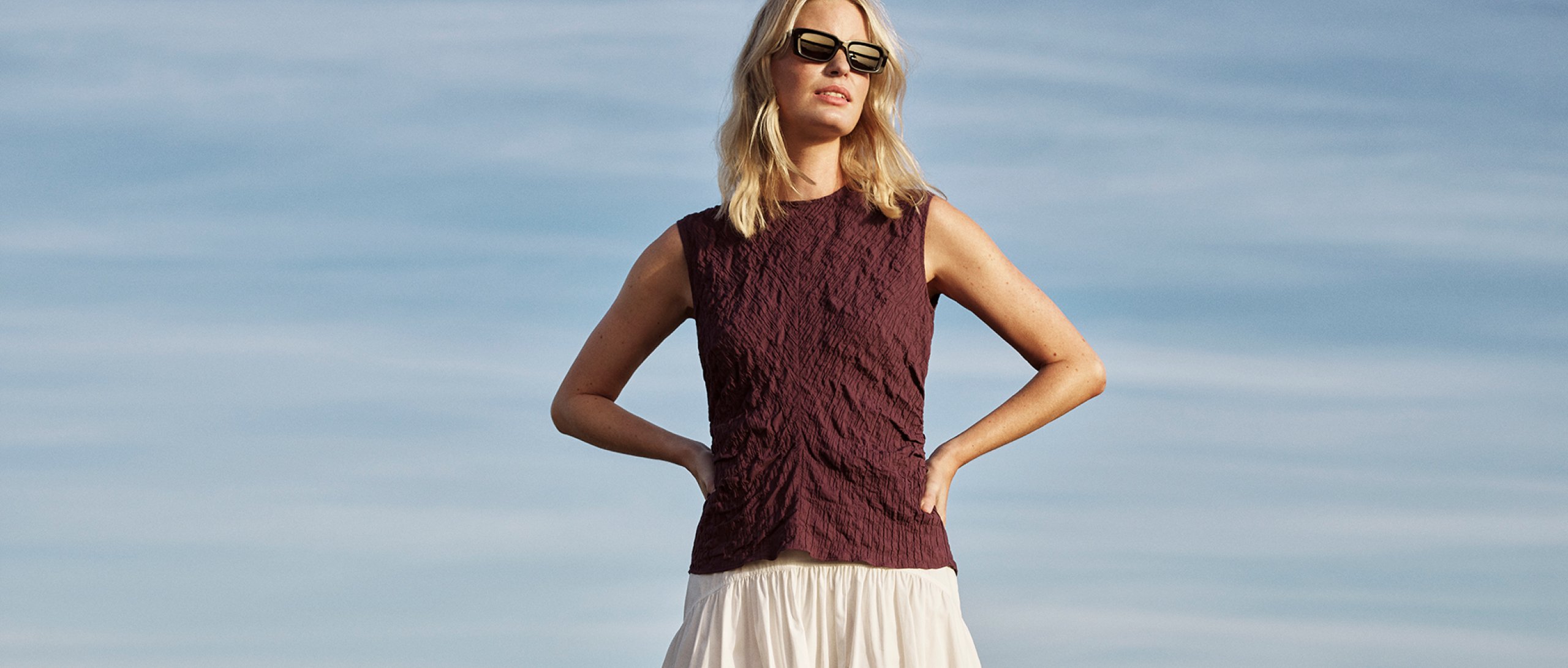 Woman wearing a sleeveless burgundy textured top with cream skirt for Whistles 15% off everything promotion