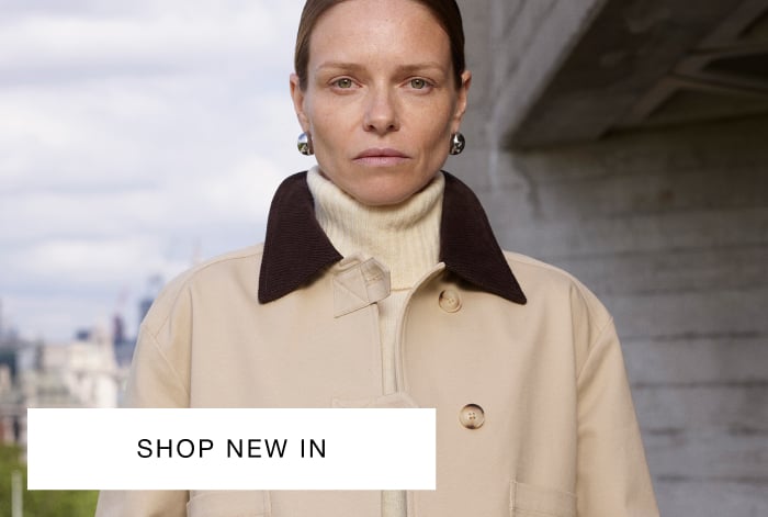 Woman wearing Whistles' cropped beige jacket with dark brown collar, cream sweater, mid-wash blue jeans, and small silver earrings
