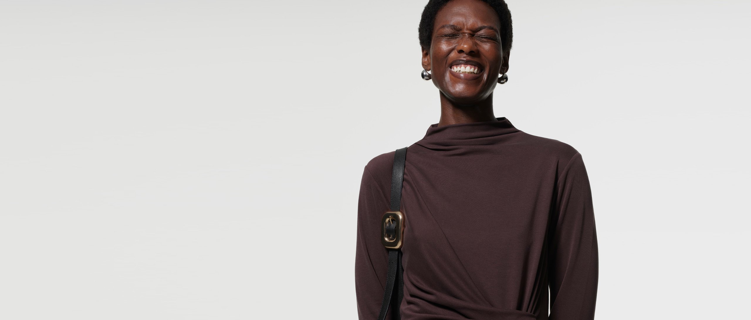 Woman wearing Whistles’ chocolate brown maxi dress with twist ruched waist and long sleeves styled with a black leather shoulder bag now 20% off.